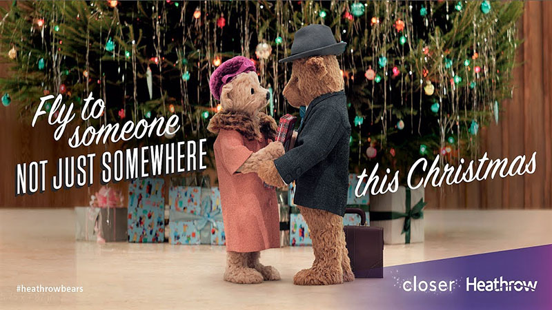 Two teddy bears -- one in a salmon coat and pink hat with a mesh veil, the other in a black coat and black fedora -- holding hands in front of a Christmas tree, with the words "Fly to someone, not just somewhere, this Christmas"