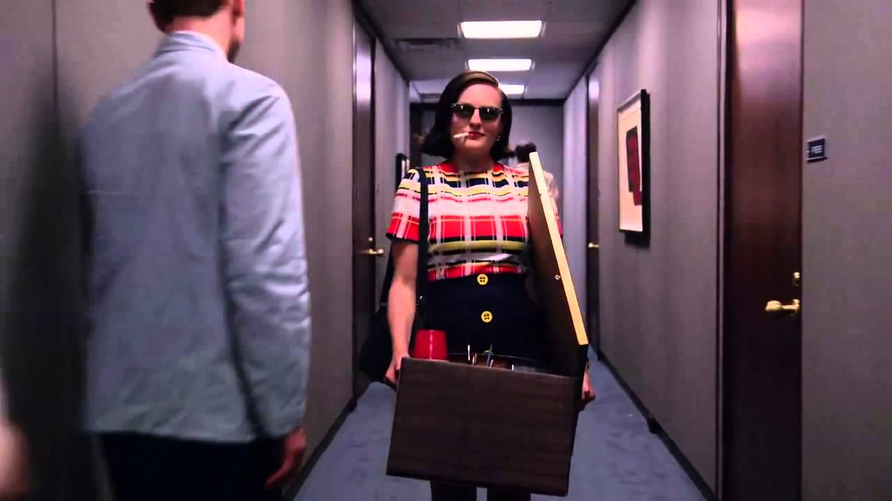 A woman ("Mad Men"'s Peggy Olson) walking down a hallway, holding a box, smoking a cigarette, and wearing sunglasses indoors