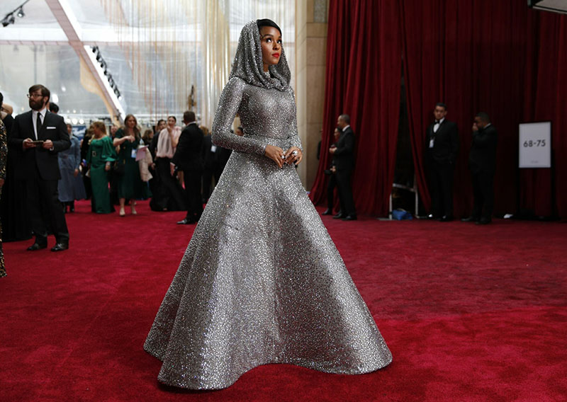 Singer Janelle Monae is a sparkly gray evening gown with a flowing hood, standing on a red carpet at the Oscars