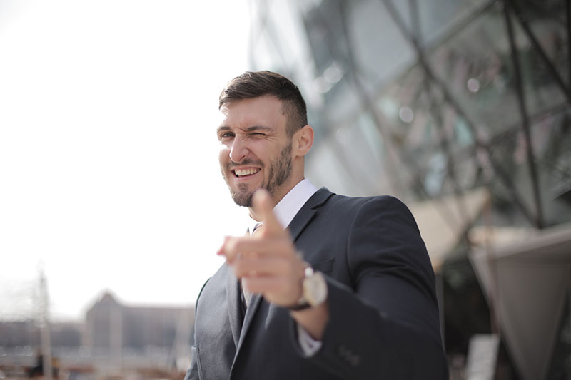 Man in black suit and white shirt, pointing at the camera and winking