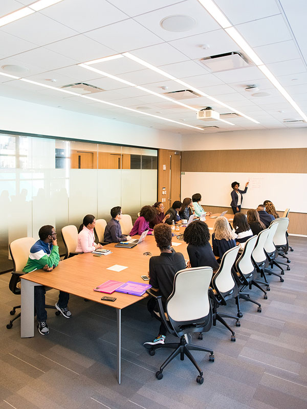 A shot of 14 people seated in beige rolling chairs around a tan wood conference table with notebook scattered on top, all on taupe faux-wood floors under a high, fluorescent-lit acoustical-tiled ceiling. At the front of the room, a woman in a dark suit points at nothing on a long whiteboard.