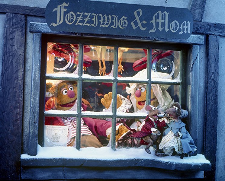 A screenshot from “The Muppet Christmas Carol,” showing a Victorian-ish storefront with a large window and a sign, in fancy type: “Fozziwig & Mom.” It’s old Fozziwig’s rubber chicken factory, as celebrated by Ebenezer Scrooge himself. Fozziwig himself (Fozzie Bear) and his mother (basically Fozzie in a Victorian dress with ruffled cuffs and a white mobcap) gaze happily out the window as two appropriately-dressed rats dance together on the sill.