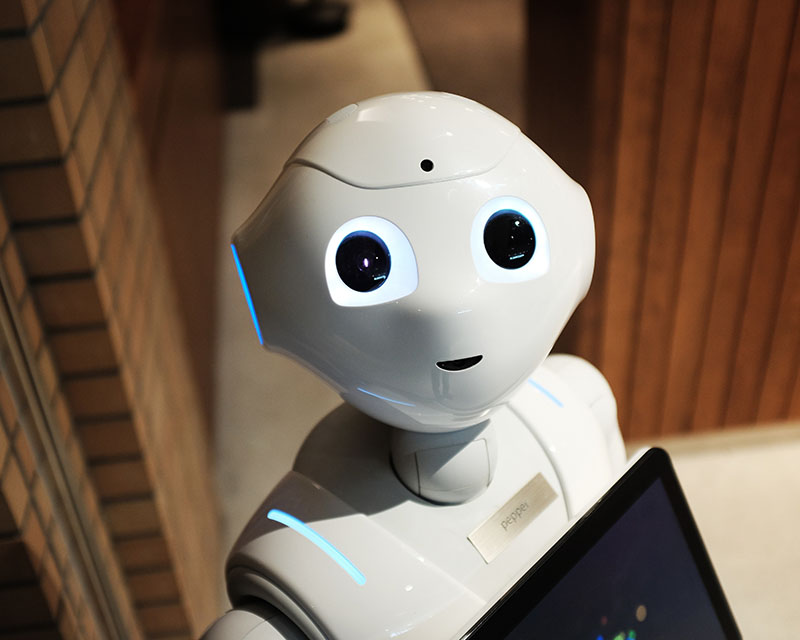 In what appears to be a wood-paneled office of some sort, we get a close-up of an adorable, shiny, white plastic robot with big, cute eyes and a li’l cute smile adorably peeking up over the edge of a laptop screen. A teeny little nameplate tells us the robot’s name is Pepper. PEPPER.