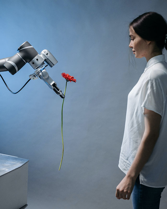 In a cold, sterile room with a blue-gray wall, a robot arm offers a long-stemmed red daisy to a woman in a white blouse and jeans. Her arms are at her sides, her back is stiff, and her face is inscrutable, but it’s apparent that whatever the robot did, they are NOT okay and she does NOT forgive him.