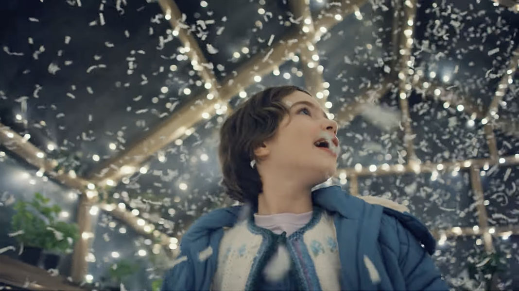 A still from Amazon’s 2022 holiday ad. A young girl with short, dark hair, wearing a blue puffer jacket, stands in the middle of a greenhouse that’s been turned into a life-sized snow globe — lights strung along the beams and a shower of shredded-paper “snow.” She’s turning around in gleeful awe, and who can blame her? It’s amazing. A human-sized snow globe would be pretty sweet in 60-degrees-in-December Alabama, that’s for damn sure.