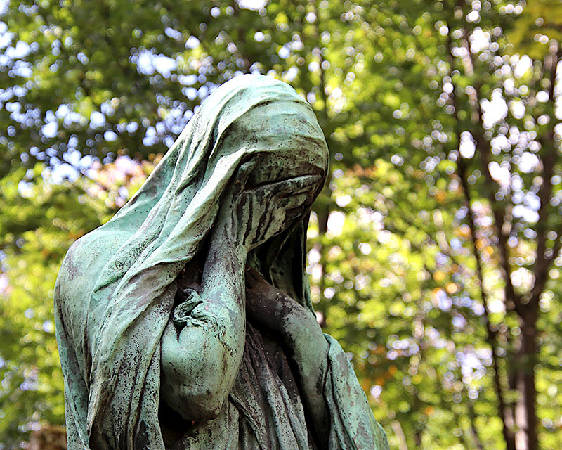 A verdigris-ed bronze statue of a woman in a veil stands against a background of trees and sunlight with her hand over her face, crying, devastated probably because her bastard client just rejected her concept for being “not really what they were looking for.”