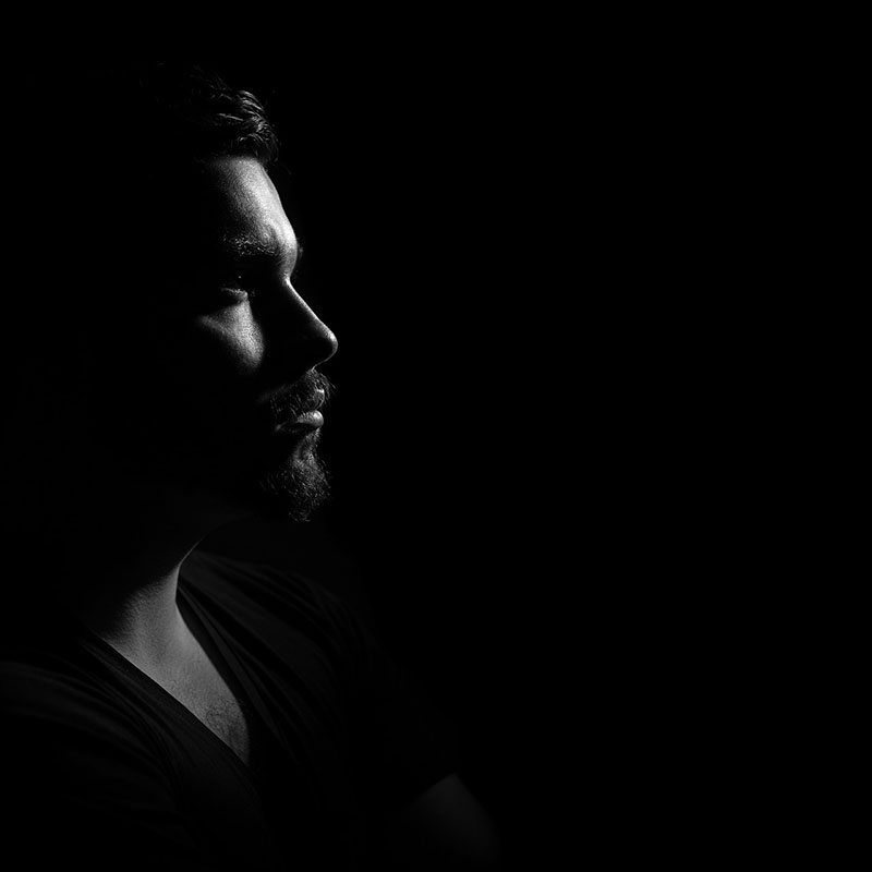 A dramatic, black-and-white profile shot of a man’s solemn face, mostly shrouded in darkness. He’s bearded and wearing a v-neck t-shirt, if that tell you anything about him.