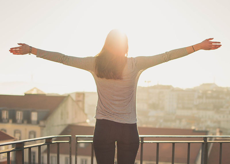 A woman stands, arms held out wide, on a balcony overlooking the rooftops of an old-and-European-looking town. She’s facing into bright sunshine that almost hides her body with its aesthetic glowiness. God, she looks happy. It’s disgusting.