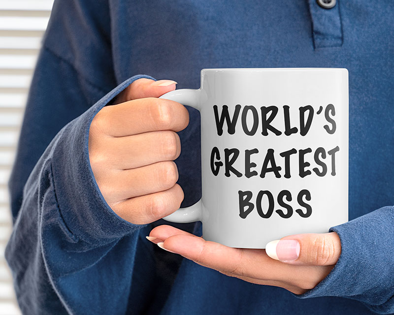 Close up of a white mug in the neatly-manicured hands of a woman in a blue blouse. The mug reads “World’s Greatest Boss” in black marker-font, and if you ask the woman if she bought it for herself, she acts REAL offended, but she doesn’t say no.