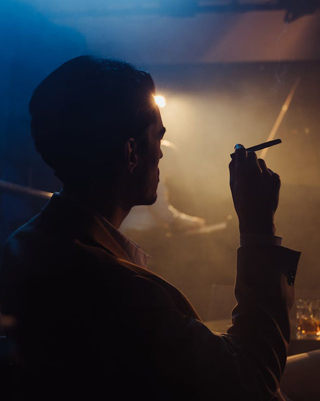 A rear 3/4 view from the waist up off a man silhouetted against a dimly lit, smoky bar, with a single spotlight casting a warm glow in the blue-toned darkness. From what we can see of the man, he’s neatly groomed and well-dressed, wearing a suit and a shirt with an open collar and smoking a cigarette, because as we all know, smoking makes you cool.