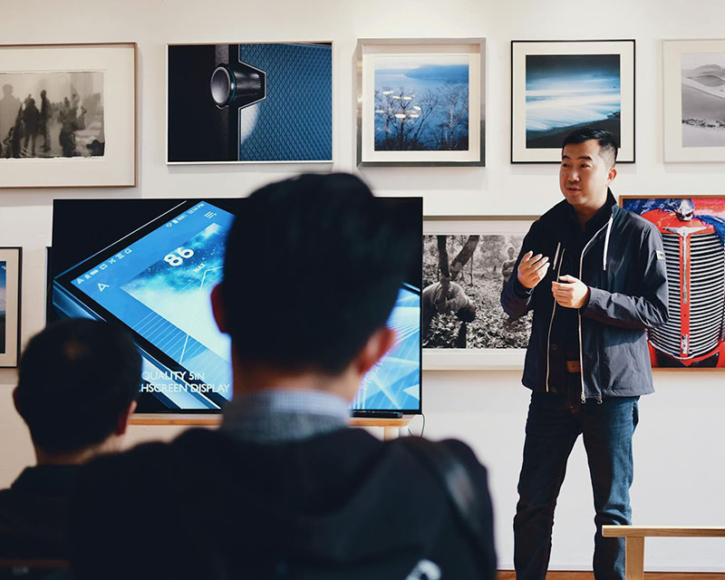 A photo of a teacher — but, like, a cool, arty teacher, middle-aged and wearing dark jeans and a navy hoodie — stands at the front of a classroom, mid-lecture. Behind him, on a white wall, are rows of framed, artsy photos. Next to him is a large screen showing what appears to be a cell phone ad. In the foreground of the photo are the backs of the heads of the students we’re sitting behind.