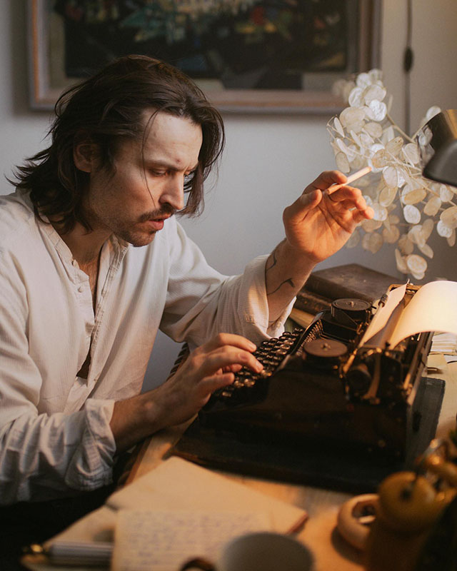 We see a man with longish, unkempt hair, wearing a half-unbuttoned loose Henley-looking top with the sleeves rolled up to reveal a tattoo on his wrist, because he’s edgy. He’s smoking a cigarette (because he’s edgy) and looking concerned as he types on an old-fashioned typewriter. On his desk, we can faintly see an open notebook, a black banker’s lamp, and a spray of white flowers.