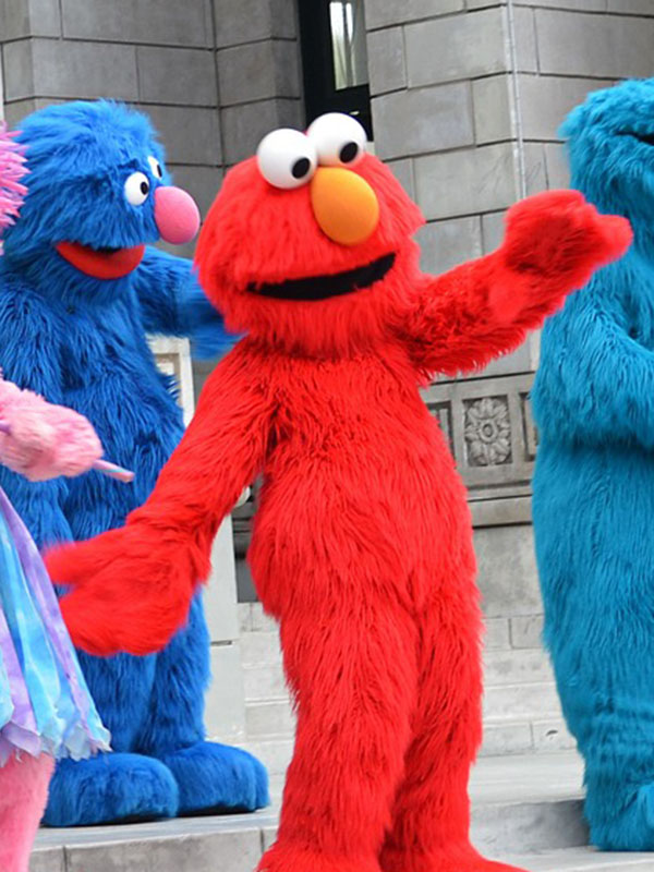 A photo of a performer costumed as Elmo, the bright red, furry Sesame Street character with a big orange nose and eyes toward the top of his head. He’s gesturing broadly, arms wide. In the background, in front of a gray stone pillar, you can see the front half of Grover, blue and furry, and the back end of Cookie Monster, turquoise and also furry.