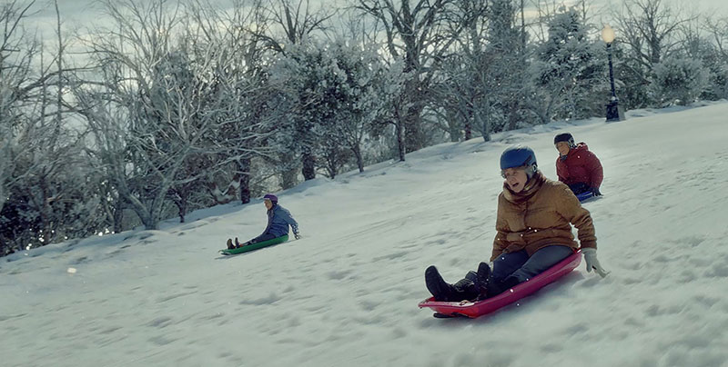 A screenshot from Amazon’s 2023 holiday ad “Joy Ride.” We see a wide shot of a snowy hill with snow-covered trees in the background. Sledding down the hill are three elderly women in hats and coats and scarves and gloves and brightly-colored plastic sleds, gleefully reliving childhood memories.