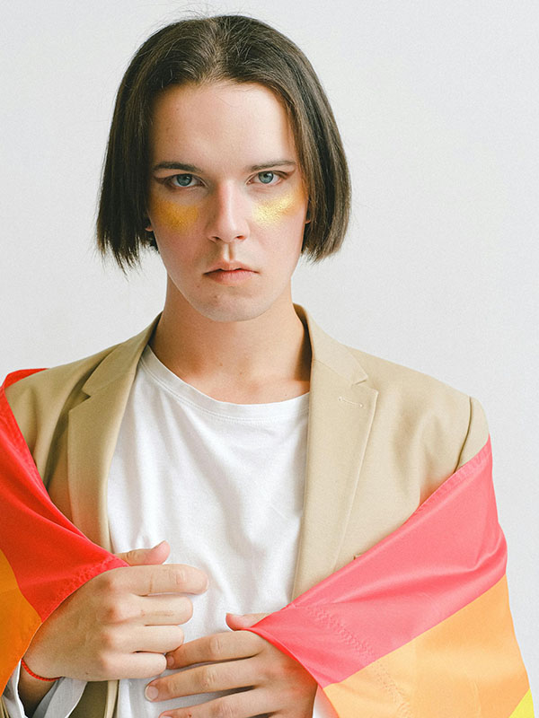 A woman with jaw-length brown hair, flat and center parted, wearing a white t-shirt and a  beige blazer, stands in front of a gray background. She has faint streaks of gold paint under her eyes, she’s wrapped in a rainbow flag, and her flat expression, looking straight at the camera, gives the impression she doesn’t want to be here.