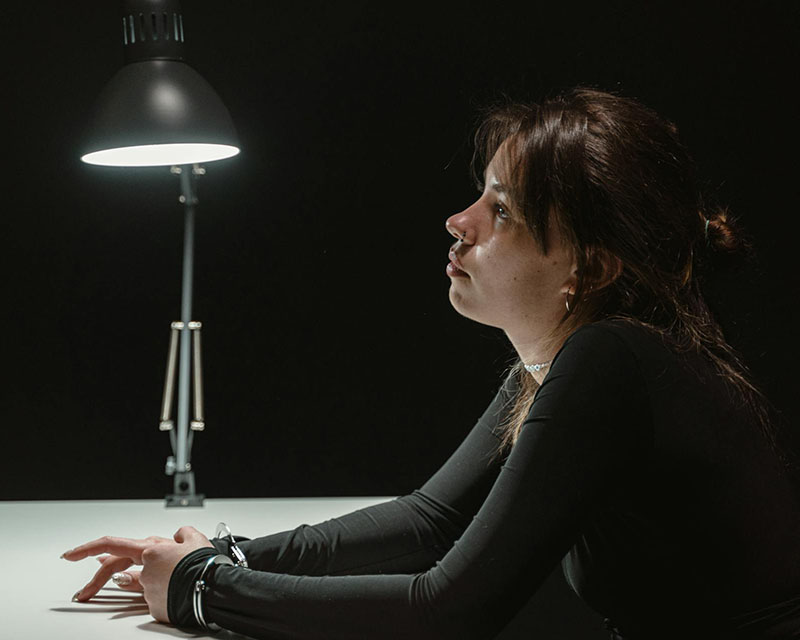 A woman with long, dark hair up in a half-ponytail and a dark gray, long-sleeved shirt sits at an interrogation table, lit only by a bright desk lamp. She’s leaning forward with her forearms on the table so you can see the handcuffs around both wrists, and her facial expression says she doesn’t talk to cops and isn’t going to tell you jack.