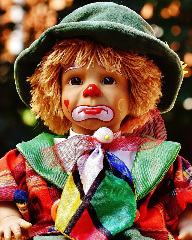 A sad clown doll sits against a blurred background. The doll has a green felt hat, yellow yarn hair, a red plaid jacket with green lapels, a harlequin-patterned necktie, and clown makeup with a li’l red nose, a red heart on one cheek, a yellow moon and star on the other cheek, and a downturned white mouth. It looks really sad, unless you’re afraid of clowns, in which case it obviously looks scary.