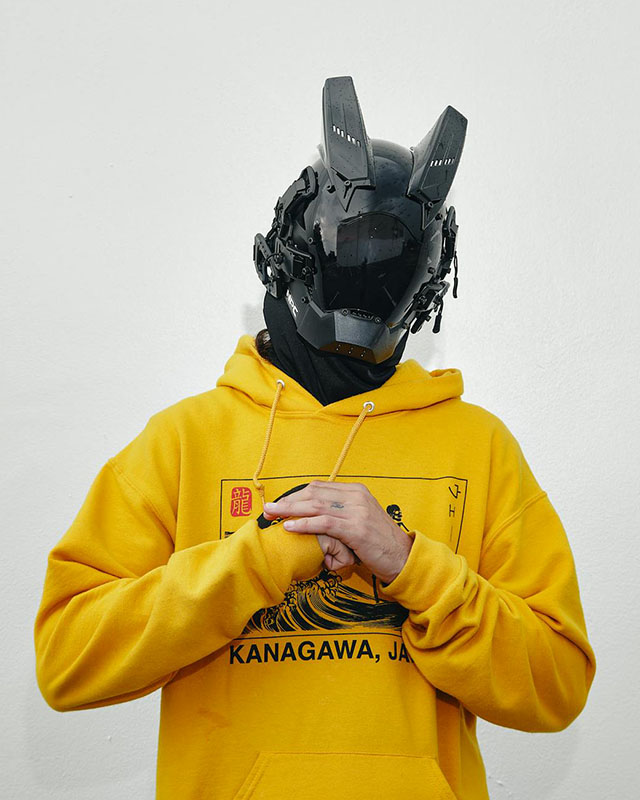 A man in a yellow hoodie stands in front of a pale gray wall, hands clasped in front of his chest. Instead of a human head, he has a robot head, black and shiny and plasticky with parts all over it and what appear to be two horns sticking out from the forehead.