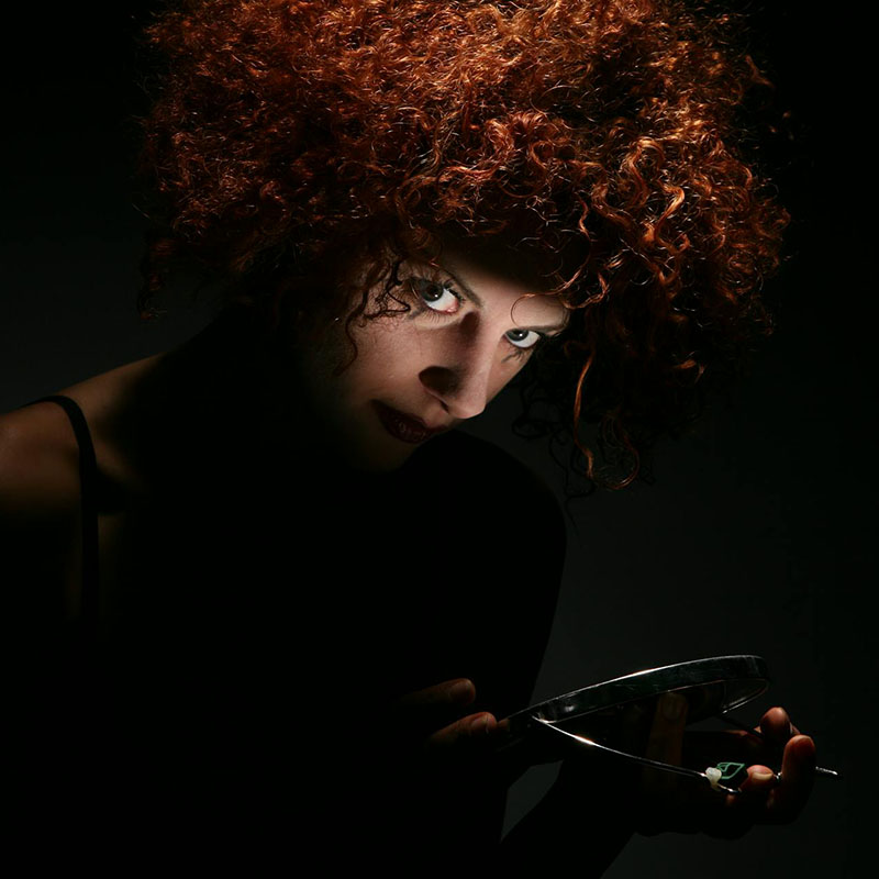 A woman with a cloud of curly, red hair stands in total darkness, angled to the right but with her eyes cut toward us. We have enough light to see her hair and her eyes and, very faintly, something silver and shiny. A switchblade? A stapler? We’d rather not find out.