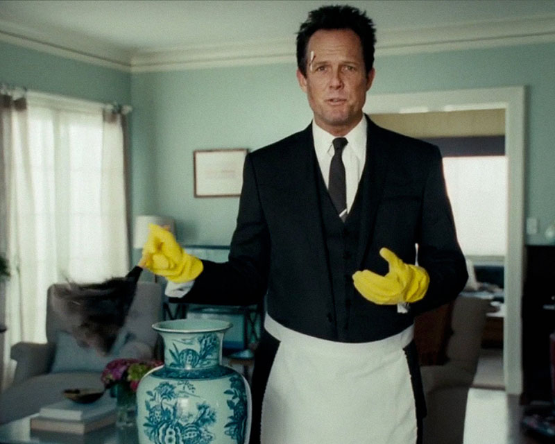 Screen shot from the Allstate “Cleaning Lady” ad. Our favorite embodiment of mayhem is dressed like a maid. He’s in a dark gray three-piece suit and tie, with a Band-aid on his forehead, along with yellow rubber gloves and a gray feather duster. He’s standing in a light blue-painted living room with comfy blue chairs and a coffee table with coffee-table books in the background, and he’s in the process of dusting an expensive-looking white and blue vase that we can just tell is not long for this world.