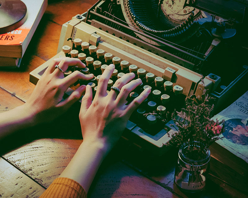 We’re looking at an old-timey typewriter sitting on a wood table, next to a small jar of dried lavender sprigs and what appears to be an old paperback romance novel. A woman’s hands are poised above the keys, ready to type something, anything, ANYTHING, as soon as the writer’s block heads out or the ayahuasca kicks in.