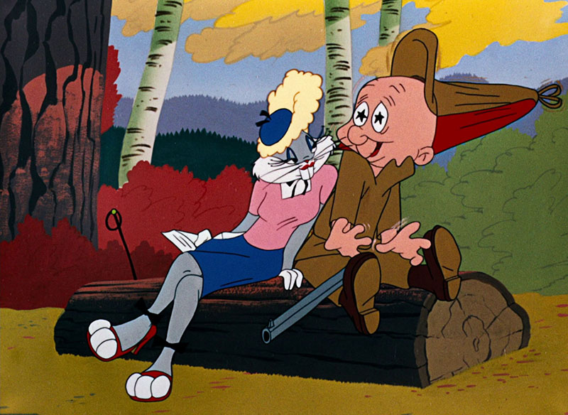 In a cel from the Looney Tunes cartoon “Rabbit Seasoning,” Bugs Bunny, in drag, next to Elmer Fudd. They’re outside with a thick brown tree, some thinner white trees with a canopy of yellow leaves, and red and green bushes in the background and, in the distance, shadowy mountains and a blue sky. They’re sitting on a brown cut log. Bugs, on the left, is in a curly blonde wig, navy blue pillbox hat, short-sleeved pink top, navy blue knee-length skirt, white wrist-length gloves, and really cute black open-toed heels with bows at the ankles. Elmer, on the right, is in a tan top and pants, dark brown boots, and a tan and red hunting cap, with a shotgun across his lap. Bugs is cuddling up close to him, all, “Hey there, big boy,” and Elmer is sitting up straight, and his pupils have turned into stars, and even his hat is standing straight up, because she may be a rabbit, and she may secretly be the boy bunny he’s been hunting all day, but at the moment, she’s a straight-up hottie.