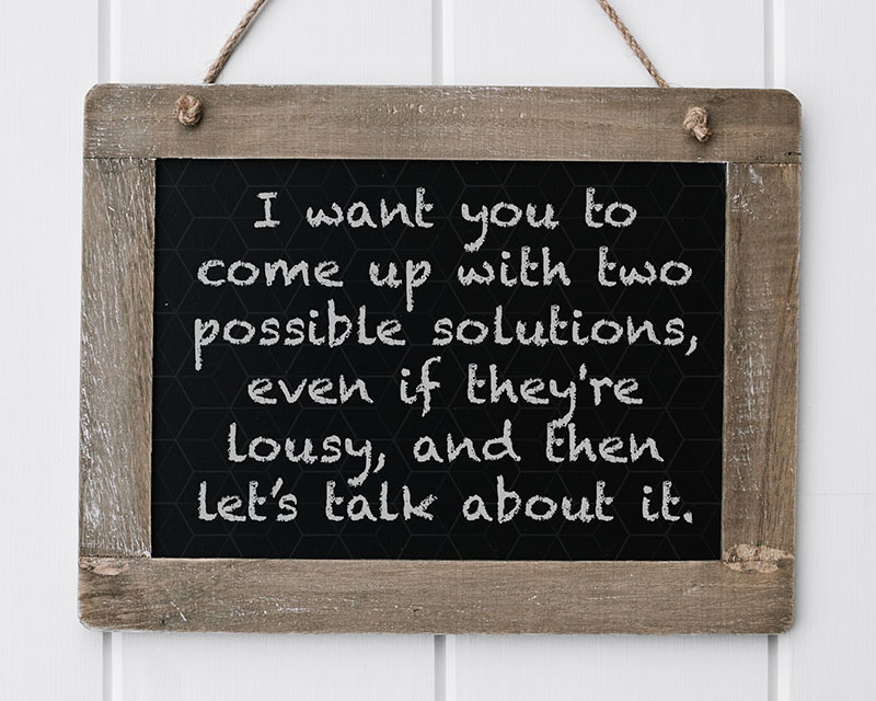 A chalkboard in a rustic wooden frame hangs by twine against a white-paneled wall. On the board, in dusty chalkboard font, is written, “I want you to come up with two possible solutions, even if they’re lousy, and then let’s talk about it.”