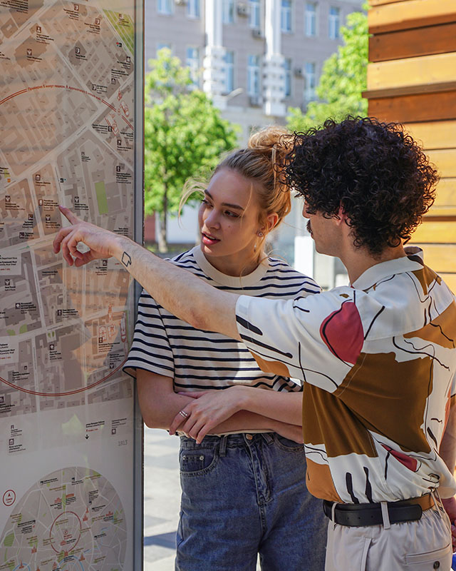 A couple stand on a city sidewalk, looking at a city map mounted behind glass on a pillar. He’s angled away from us, such that we can see his full head of tight, dark curls, his dark mustache, and his Picasso-looking short-sleeved cream and brown and red collared button-down tucked into his rumpled khakis. She’s got her blonde haired piled on top of her head in a messy bun and is wearing a black-and-white-striped short-sleeved shirt tucked into stone-washed, high-waisted ‘80s jeans. He’s pointing at a spot on the map, and the expression on her face tells us that whatever he’s pointing at, she’s not impressed.