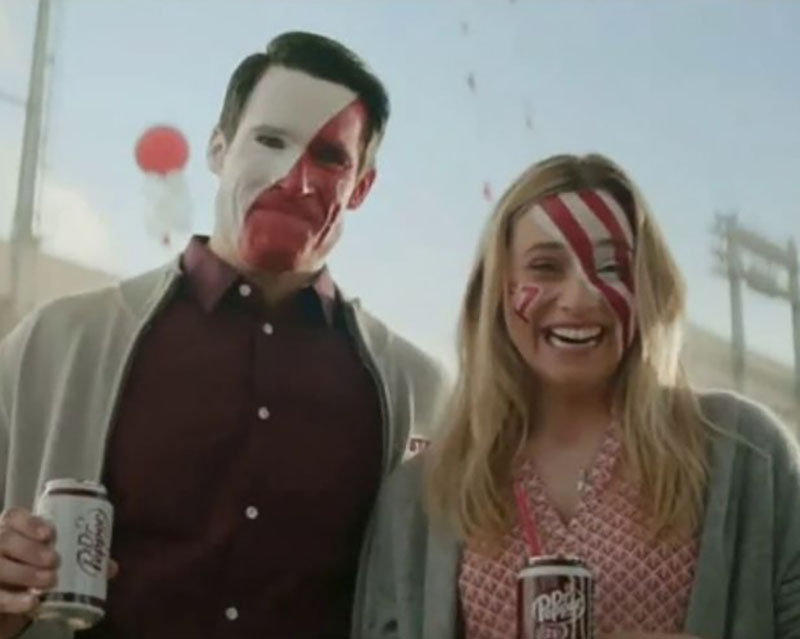 Screen shot from the Dr. Pepper “Fansville: First Words” ad. A couple stands silhouetted against a blue sky and a football stadium in the background. His dark hair is parted neatly on the side, and he’s wearing a gray zip cardigan, a burgundy button-down shirt, and a full face of paint, dividing his face diagonally into a white half and a red half, and he’s holding a can of Diet Dr. Pepper. She’s also wearing a gray zip cardigan, with a red gingham v-neck top, a face painted in red and white stripes and a red #1, and a can of Dr. Pepper. They’re both smiling, but her smile is about as stiff as you’d expect from a woman whose husband is about to find out their Tech-loving baby might not actually be his.