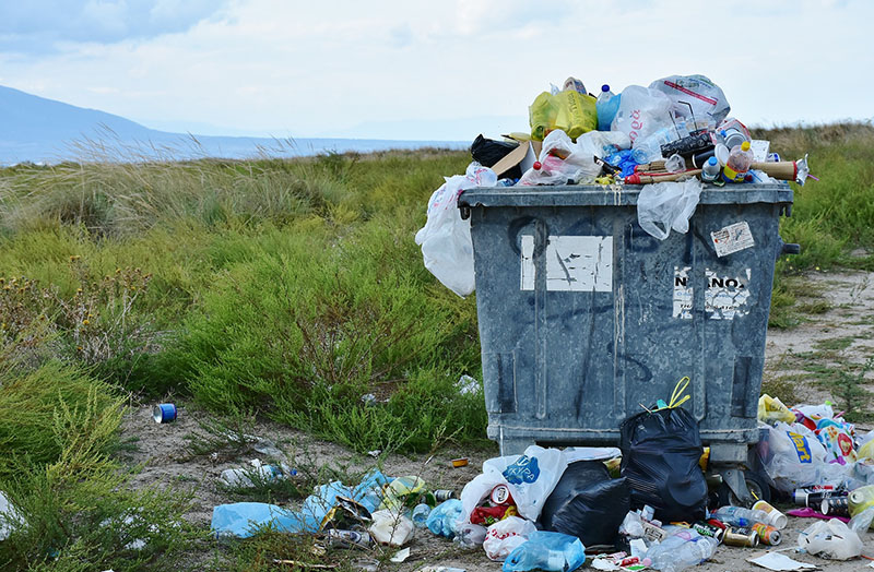 A faded gray dumpster overflowing with trash, and surrounded by trash, at the edge of a field of patchy scrub grass and low-lying plants and bare dirt, with a mountain and hazy blue sky with just a few fluffy clouds in the distance. So oddly pretty, but still garbage.