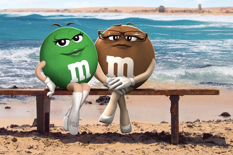 The newly redesigned green and brown M&M mascots sit on a wooden bench on a sandy beach, with a blue sky and blue waves topped with white foam in the background. Both are facing the camera with their bodies angled toward each other, and the green one, sitting on the left, is smiling slyly. The brown one, sitting on the right, wearing glasses and white pumps, has a neutral expression. The green M&M is wearing white knee boots and has one hand on her hip, her ankles crossed. The brown one has her legs crossed at the knees. The green M&M has one hand on the brown one’s knee, and the brown one’s hands are clasped on top of the green one’s.