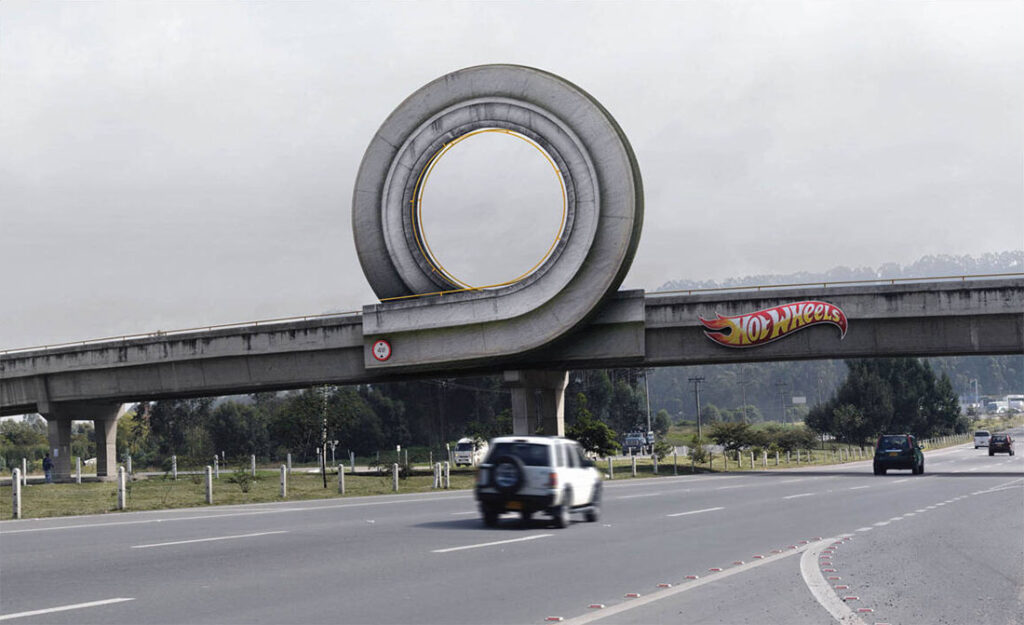 A photo of a highway overpass showing a 2011 outdoor board in Colombia for Hot Wheels by agency Ogilvy & Mather. In the background, we can see that the day is gray and foggy, as cars drive on a six-lane highway with a grassy, tree-lined median in the center. A steel and concrete overpass spans the highway, but right above the median is a big loop, making the overpass look like a life-sized version of the kind of looping track you might have played with your Hot Wheels toy on when you were a kid. To the right of the loop is the Hot Wheels logo in white and yellow letters on a red flame background. It’s badass.