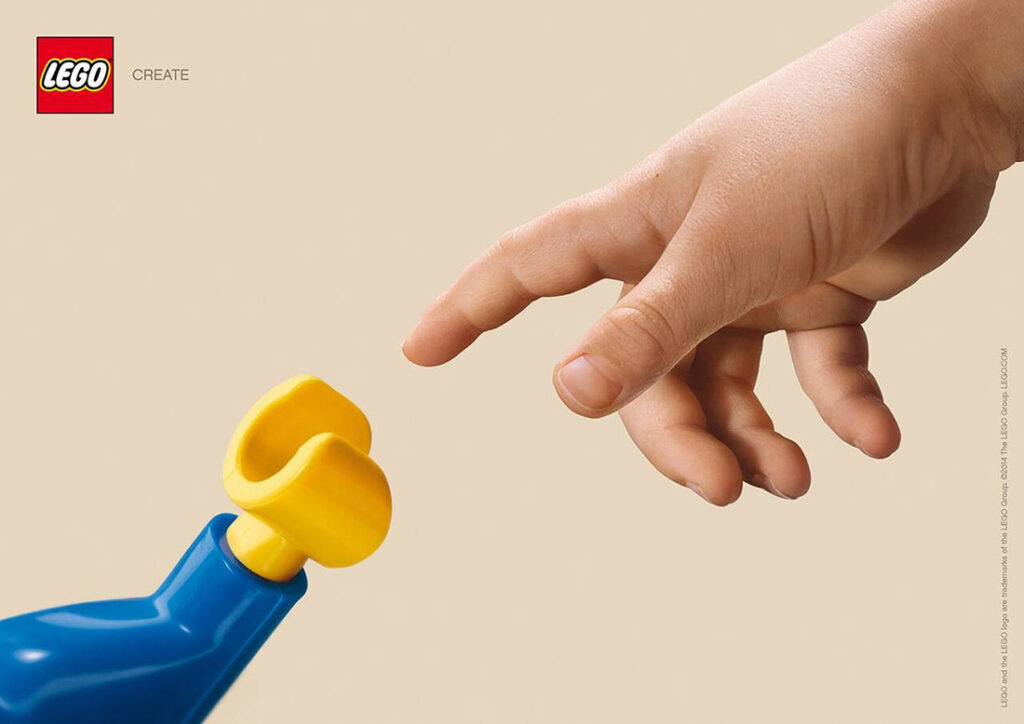 A 2014 Lego ad by agency Jung von Matt. Against a cream-colored background, a white man’s hand reaches out of the upper right-hand corner, one finger extended like Michelangelo’s “The Creation of Adam.” But the hand reaching up from the lower left-hand corner to touch it is a yellow plastic Lego figurine hand in a blue plastic shirtsleeve. In the upper left-hand corner is the Lego logo and the word “create.”