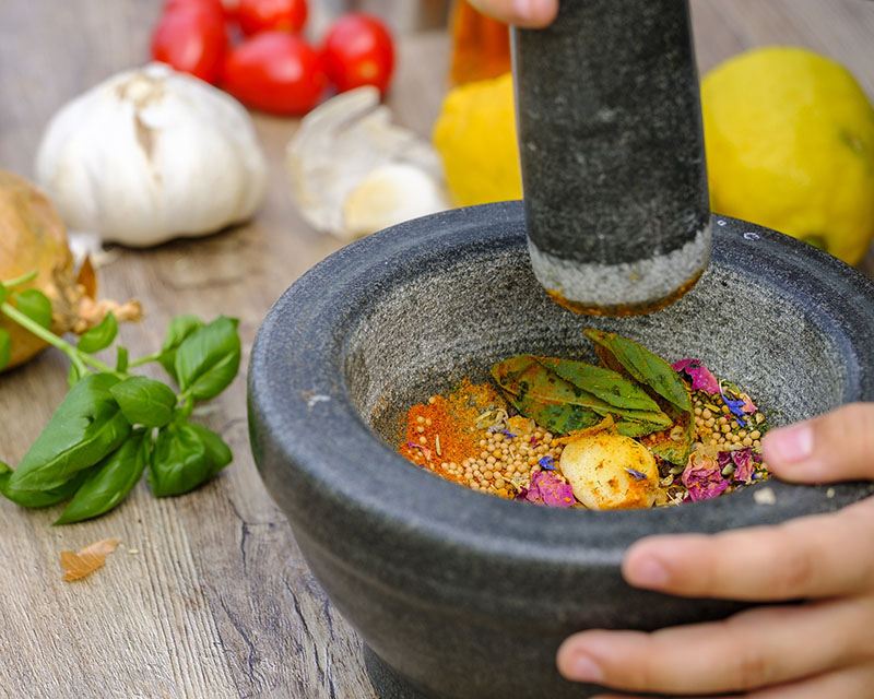 A close-up of a gray stone mortar and pestle, wielded by two hands. The cooking tools sit on a wood table in front of basil leaves and a garlic cloves and a few tomatoes, and it the bowl is full of leaves and flowers and seeds, all pretty and all in the process of getting violently ground up.