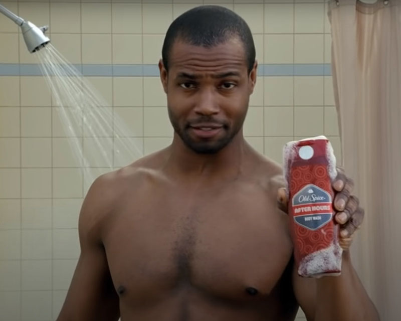 Screen shot from the Old Spice “The Man Your Man Could Smell Like” ad. A handsome, well-tended man in a toned pair of pecs and not much else stands in a yellow-tiled bathroom with a running shower head and beige shower curtain in the background. He’s holding up a red bottle of Old Spice After Hours body wash, and absolutely no one is focused on the bottle.