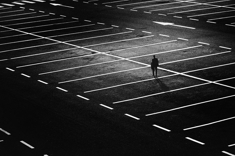 A black-and-white photo of an empty parking lot at night with one guy silhouetted in the distance, standing in a space, all alone, so very alone, always alone.