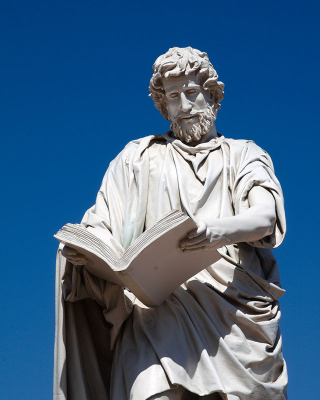 A white-gray marble statue stands against a brilliantly blue sky — a bearded man with curly hair, wearing flowing robes, holding a large, open book at arm’s length. What book is it? What is he thinking about it? It’s probably the concept he came up with that just got rejected by the client, but he doesn’t care because that. Concept. Was. Fire.