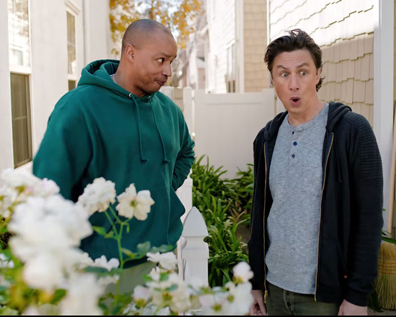 Screen shot from the T-Mobile “Musical: $50” ad featuring Donald Faison and Zach Braff from “Scrubs.” The two are standing in a breezeway of a house clad in beige shingled siding, behind a profusion of white flowers. Donald, wearing a teal hoodie and khaki cargo pants, is looking at Zach, kind of pinched. Zach, wearing a gray Henley, a zip hoodie, and jeans, looks into the middle distance, apparently shocked. At what? I don’t remember. It’s been a while since I watched the ad.