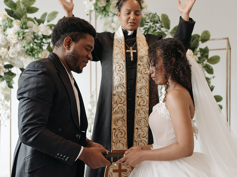 A waist-up shot of a bride and groom, with a minister in the background in a black robe, gold sash, and gold cross, hands raised in blessing. Behind her, the front of the church has a white wall with white flowers and greenery cascading down. On the left, the groom, who has dark skin and close-cropped hair, wears a black-and-white tuxedo. On the right, the bride, who has dark skin and long, curly hair, is wearing an ivory strapless dress with lace trim and a full skirt, and a tulle veil pinned to the back of her hair and trailing downward and out of the frame. They’re both looking downward and holding a Bible together. They’re so pretty. Definitely someone is crying at that wedding.