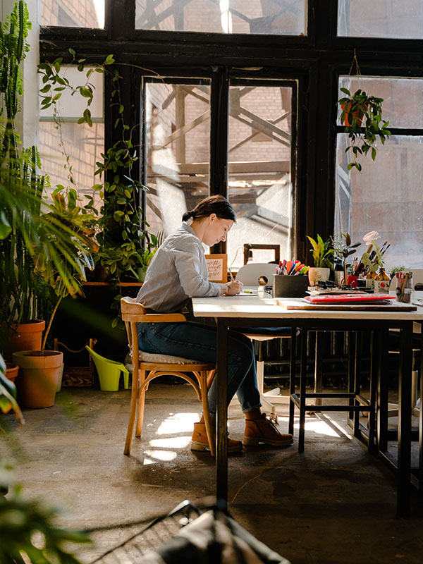 A woman in a gray sweatshirt, jeans, and tan work boots sits at a table in what appears to be the sunroom of an older building, with big windows framed by dark wood. The table is cluttered with pen cups, folders, and potted plants, and she’s writing. The entire room is filled with plants, in fact — potted ones, hanging ones, climbing ones, big ones sitting on the floor, huge ones reaching nearly to the ceiling. The woman is clearly into writing, and growing things.