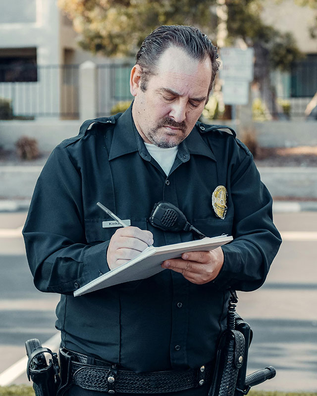 Photograph of a middle-aged police officer with slick-backed hair and a goatee on a downtown sidewalk, wearing the classic navy-blue uniform and utility belt and badge and body cam, writing on a notepad.