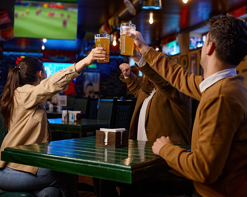 Three friends sit at a sports bar, around a square table. The two men are wearing brown sweaters, and the woman is wearing a light-brown blouse. They’re facing away from the camera, looking at a big TV screen at the back of the room and toasting with pints of beer.