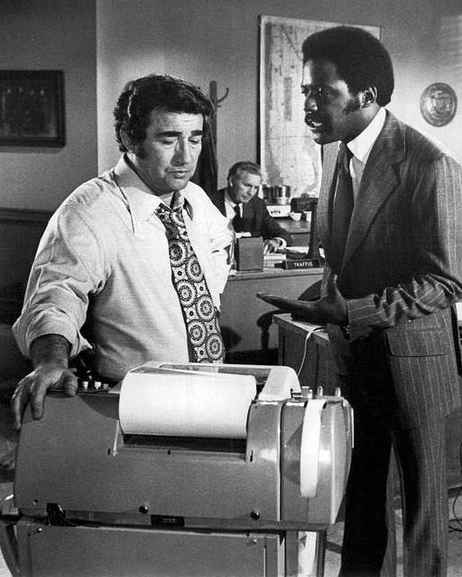 A black-and-white screenshot from the “Shaft” TV show. Shaft, a dark-skinned black man with a short afro and a mustache, wearing a dark pinstriped suit, stands next to a shorter white guy with a nondescript hairdo in a white shirt and patterned tie next to… a machine, I don’t know what, it looks like an adding machine if it were the size of a fax machine, it was the ‘70s. The black guy looks emphatic about something, but the white guy doesn’t seem concerned.