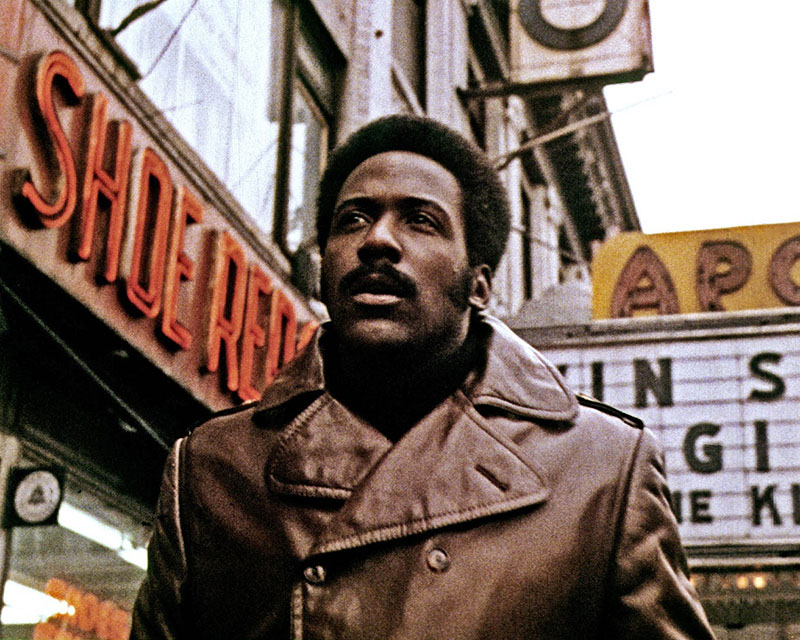 A screenshot from the movie “Shaft.” We get a chest-up view of Shaft, a dark-skinned black man with a short afro and a mustache, wearing an awesome brown leather blazer, as he walks down a city street past a building with “Shoe Repair” in lights on the wall. Behind him, we can see just a little bit of a movie theater marquee.