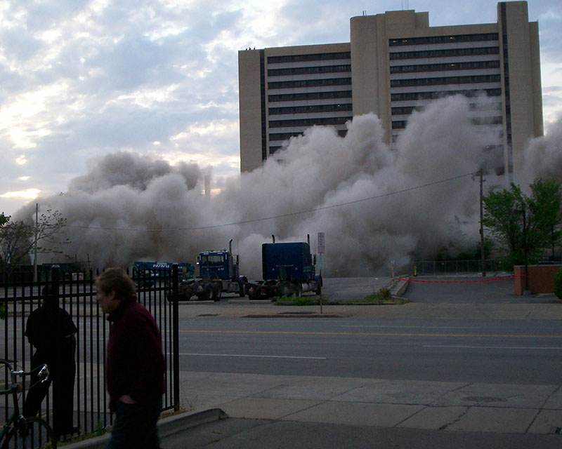 Photo taken from a safe distance of an office building being imploded on a sunny day, with clouds of smoke and dust billowing around the base as the building begins to slightly tilt. In the lower corner of the shot, two men stand, unimpressed.