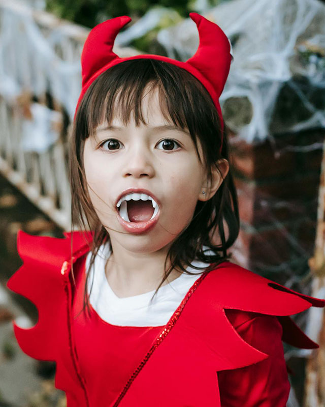 Chest-up photo of a young, brown-haired girl, maybe five, in a red devil costume — a little red top with a pointy collar and a red devil-horns headband, with her mouth wide open to show off white plastic fangs. She is adorable.