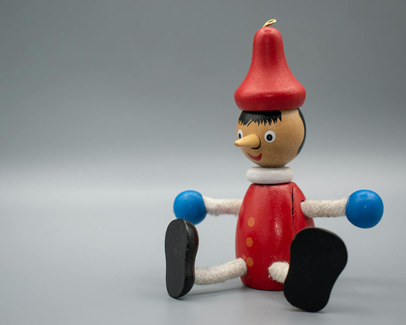 Shot of a wooden Pinocchio toy with a red hat and body and blue hands and, of course, a long nose, sitting at the front right of the frame against a gray, shadowed background.