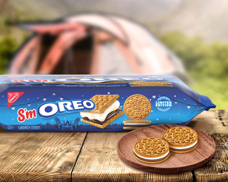 A photo showing a side view of a box of s’mores-flavored Oreos — S’mOreos, if you will. They’re sitting on a rough wood picnic table next to a wooden plate with two S’mOreos on it, with a blurry tent and outdoor setting in the background.