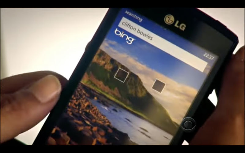 Screenshot from an episode of “Hawaii Five-0.” We’re looking at the screen of a black LG phone held in a woman’s hand against an off-white background. On the screen is a lovely shot of a green mountain against a blue sky with white clouds, at the edge of a cay in Hawaii with pink flowers in the foreground. At the top of the screen is a white search bar with the words “clifton bowles” in it, and immediately below that is the white Bing logo.