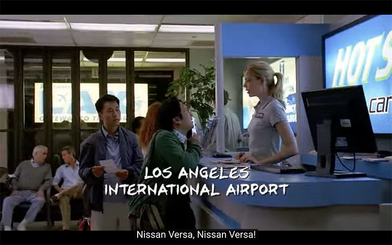 Screenshot from “Heroes.” Hiro Nakamura and Ando Masahashi, two young Japanese men in button-down shirts and cardigans, stand at an airport car rental counter, talking to a young blonde woman in a light blue polo shirt. A caption in comic book-style font reads, “Los Angeles International Airport,” and the closed caption at the very bottom reads, “Nissan Versa, Nissan Versa!”